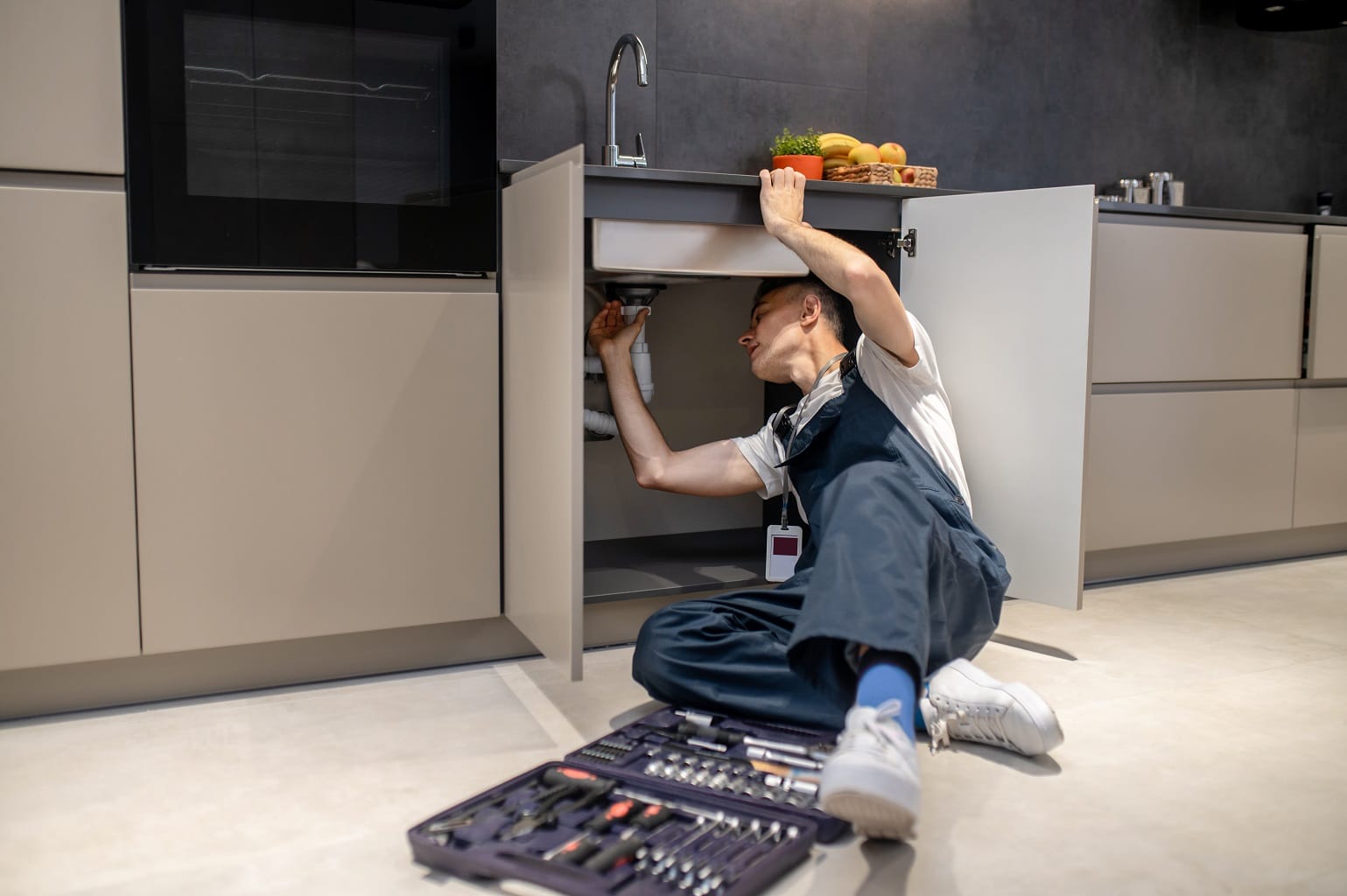Household repair. Middle aged man in blue jumpsuit reclining professionally inspecting pipe touching hand under sink in stylish modern kitchen